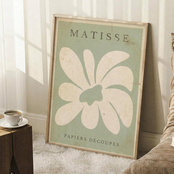 May include: Framed vintage-style art print with sage green background featuring large cream-colored abstract flower or leaf cutout design, displayed leaning against white wall corner on textured rug, with coffee cup on wooden block and beige throw blanket visible in cozy room setting with natural window light.