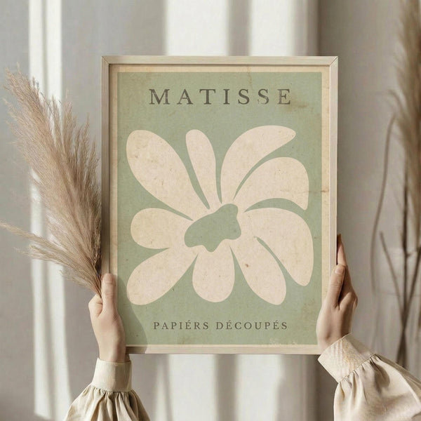 May include: Person in cream blouse holding framed art print featuring large white abstract flower shape on sage green background with "MATISSE" and "PAPIERS DÉCOUPÉS" text, shown with dried pampas grass against white curtained window in natural lighting