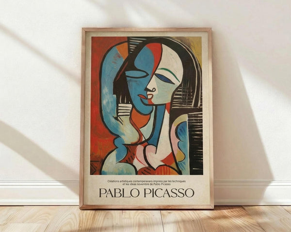 May include: Framed Pablo Picasso-style abstract portrait poster leaning against white textured wall on light wooden floor. Artwork features cubist woman with split blue and white face, black hair, red lips, geometric shapes in vibrant colors including orange, red, and yellow background. Natural light casts soft shadows across the scene. Light wood frame surrounds the vintage-style art print with cream matting and artist name below image.