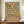 May include: Framed vintage-style botanical poster with cream background featuring stylized green and blue leaves, orange sun, and earth-tone floral motifs surrounding "Nature's Essence" text. Brown wooden frame displayed on walnut credenza beside white ceramic vase and stacked books against beige wainscoted wall in modern home interior.