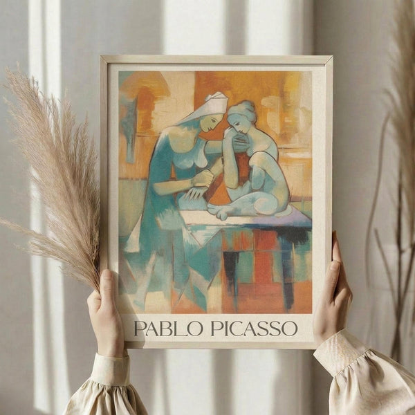 May include: Person in cream ruffled sleeves holding framed Picasso-style art print featuring two seated figures in blue and orange cubist style, alongside dried wheat stems against white curtained background. Light wood frame with artist name at bottom.