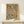 May include: Framed vintage-style botanical poster with earth-toned leaves, branches, and circular elements in greens, browns, and rust colors on cream background. Wood frame sits on light wooden surface against white wall beside small white wrapped gift with twine and greenery sprig.