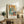 May include: Framed Pablo Picasso exhibition poster featuring two abstract cubist figures in blue and orange geometric forms, displayed on rustic wooden sideboard with books, ceramic vase, fern plant, and cream upholstered furniture in warm neutral living room setting.