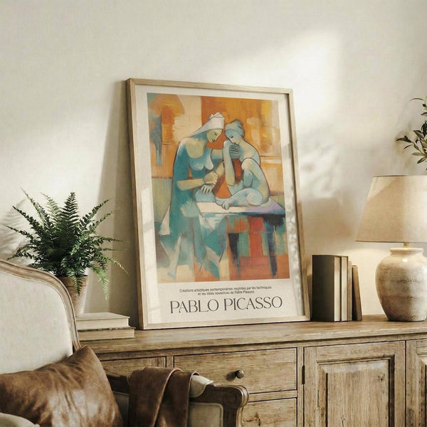 May include: Framed Pablo Picasso exhibition poster featuring two abstract cubist figures in blue and orange geometric forms, displayed on rustic wooden sideboard with books, ceramic vase, fern plant, and cream upholstered furniture in warm neutral living room setting.