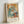 May include: Framed Pablo Picasso exhibition poster featuring two women in cubist style with soft blue, orange and beige tones, displayed in light wooden frame against white wall. The artwork shows geometric, angular figures seated at a table with French text below identifying the artist.