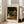 May include: Framed Claude Monet exhibition poster featuring a woman in white dress walking through a blooming garden with flowering trees. The vintage-style print has cream matting and light wood frame, displayed on wooden floors next to a beige upholstered chair in a bright, sunlit room with white walls and large windows.