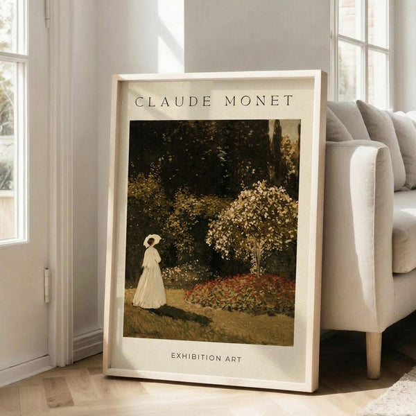 May include: Framed Claude Monet exhibition poster featuring a woman in white dress walking through a blooming garden with flowering trees. The vintage-style print has cream matting and light wood frame, displayed on wooden floors next to a beige upholstered chair in a bright, sunlit room with white walls and large windows.