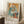 May include: Framed Picasso-style cubist artwork showing two abstract female figures in blue, orange, and cream tones, leaning against white wall near wooden side table with coffee cup, displayed on textured white rug with beige throw blanket visible.