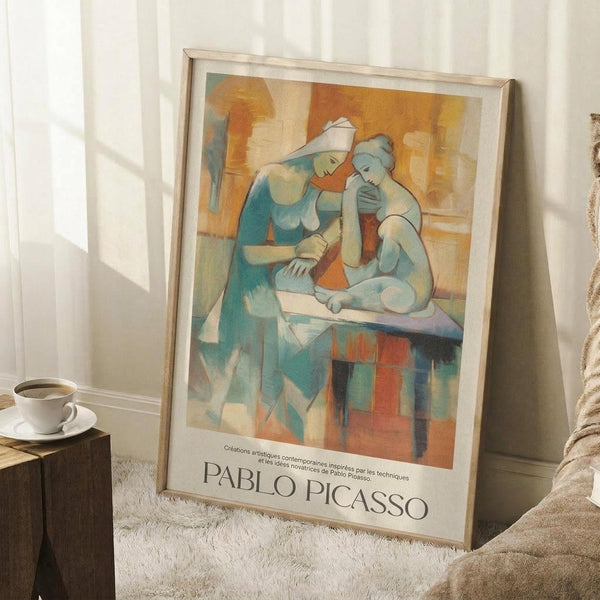 May include: Framed Picasso-style cubist artwork showing two abstract female figures in blue, orange, and cream tones, leaning against white wall near wooden side table with coffee cup, displayed on textured white rug with beige throw blanket visible.