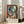 May include: A framed Picasso-style cubist portrait poster featuring a woman with geometric blue and white facial features, red lips, and flowing hair rendered in bold colors including orange, red, and black. The artwork displays French text and "PABLO PICASSO" at the bottom. The cream-colored frame leans against a white wall next to a beige sofa in a bright, modern living room with large windows and natural lighting.