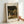 May include: Framed art print of Claude Monet painting showing woman in white dress with parasol walking through garden path surrounded by blooming trees and colorful flower beds. Print has cream matting with "CLAUDE MONET" and "EXHIBITION ART" text, displayed in light wood frame against white wall with hand holding corner for scale.