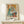May include: Framed Pablo Picasso exhibition poster showing two abstract figures in blue, orange and cream cubist style, displayed on light wood console table with small wrapped gift box beside it against cream wall