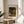 May include: Framed Claude Monet exhibition art print displayed on light wood round side table next to beige upholstered chair. Print shows woman in white dress with parasol walking through blooming garden with flowering trees. Warm sunlight streams across neutral-toned room with plant leaves visible in corner. Thin gold or brass frame with cream matting.