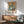May include: Framed Pablo Picasso poster featuring two abstract figures in cubist style with blue, orange, and beige geometric forms, displayed above a mid-century modern wooden credenza with white ceramic lamp. Light green upholstered armchair with wooden frame sits nearby in bright, minimalist room with white walls and natural sunlight creating shadows.