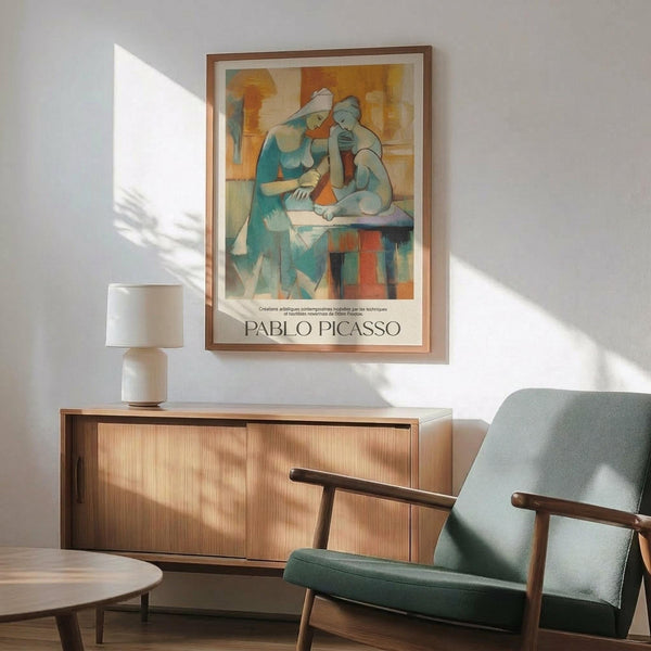 May include: Framed Pablo Picasso poster featuring two abstract figures in cubist style with blue, orange, and beige geometric forms, displayed above a mid-century modern wooden credenza with white ceramic lamp. Light green upholstered armchair with wooden frame sits nearby in bright, minimalist room with white walls and natural sunlight creating shadows.