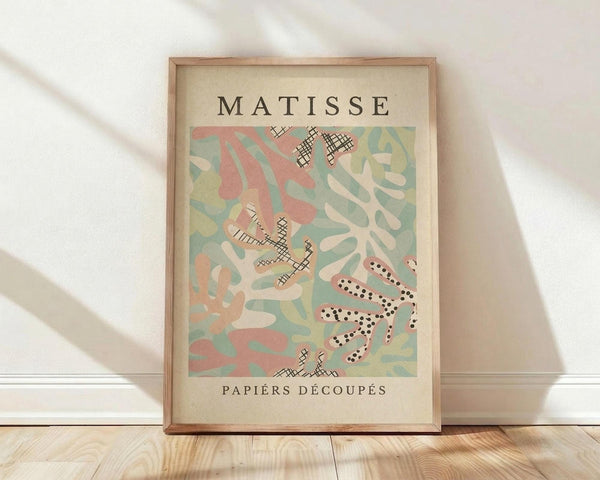 May include: Framed vintage-style art print featuring abstract cut-out shapes in soft coral pink, sage green, cream and beige colors with crosshatched and dotted patterns. The poster displays "MATISSE" at top and "PAPIERS DÉCOUPÉS" at bottom in black text on cream background. Natural wood frame sits on light wooden floor against white wall with soft window light casting shadows across the scene.