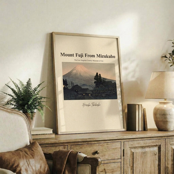 May include: Framed vintage-style poster showing Mount Fuji at sunset with pink sky and dark silhouetted landscape, displayed on rustic wooden sideboard alongside fern plant, books, ceramic vase and table lamp in warm-toned living room setting