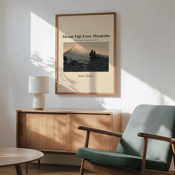 May include: Framed vintage Japanese woodblock print of Mount Fuji at sunset hanging on white wall above wooden sideboard. Print shows mountain with pink-orange peak, dark foreground landscape, and cream matting in thin brown frame. Modern living room setting with mint green upholstered chair, round wooden coffee table, and ceramic table lamp with white shade. Natural sunlight creates diagonal shadows across the scene.