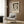 May include: Paul Klee exhibition poster featuring abstract figure with angular face and crescent moon in muted tones, displayed in light wooden frame on textured wood wall. Modern beige armchair with geometric patterned pillow sits below, alongside brass floor lamp with white marble base and open book on dark side table.