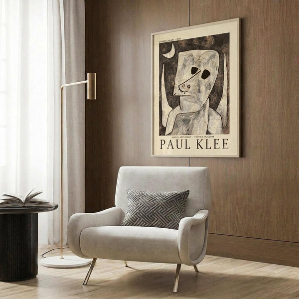 May include: Paul Klee exhibition poster featuring abstract figure with angular face and crescent moon in muted tones, displayed in light wooden frame on textured wood wall. Modern beige armchair with geometric patterned pillow sits below, alongside brass floor lamp with white marble base and open book on dark side table.