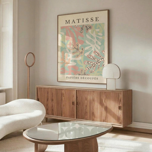 May include: Framed Matisse art print displayed above wooden sideboard in modern living room. Poster features colorful abstract cut-out shapes in pink, green, and white with black polka dot patterns. Room shows cream textured chair, glass oval coffee table, white table lamp, and neutral walls with natural lighting.