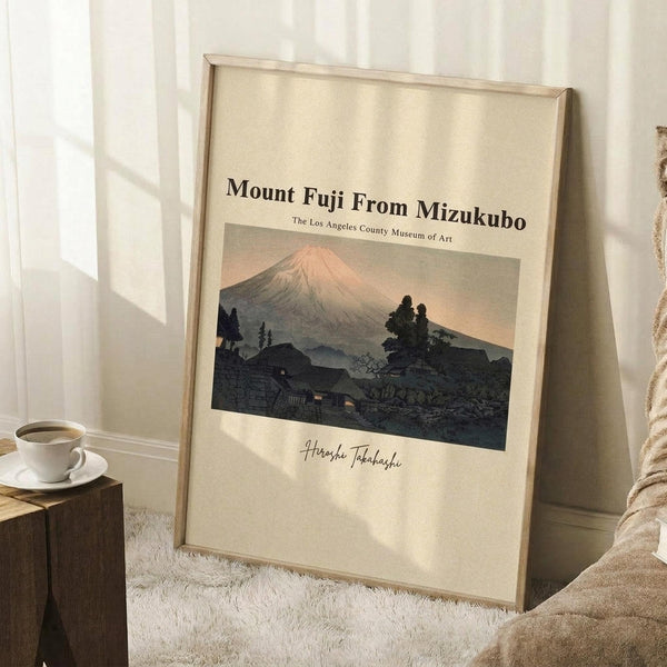 May include: Framed vintage-style poster print of Mount Fuji with pink summit and dark silhouetted village buildings and trees in foreground, displayed in light wooden frame against white wall corner. Scene shows cozy room setting with cream textured rug, wooden side table with white coffee cup and saucer, and beige blanket. Natural sunlight creates soft shadows across the cream-colored poster background with museum attribution text and artist signature visible below the Japanese landscape artwo