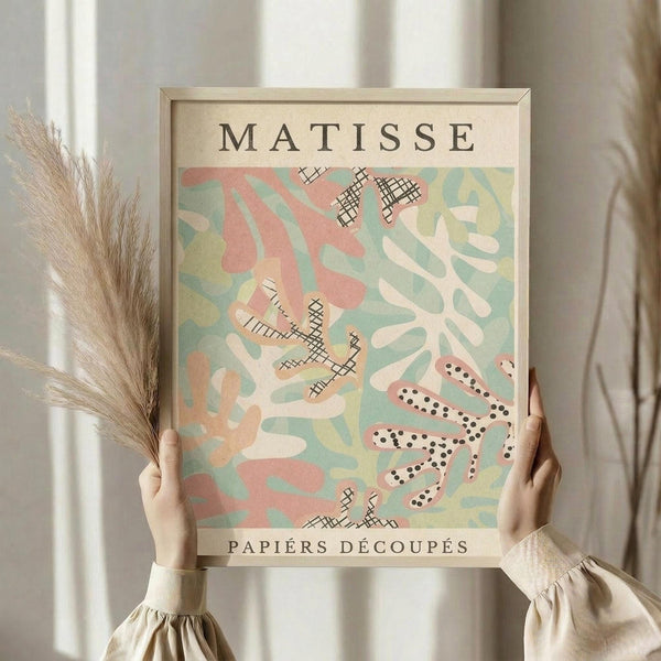 May include: Person holding framed art print in light wood frame against white curtained window. Print features abstract cut-out shapes in soft pink, sage green, cream and yellow with black crosshatch patterns and dotted snake-like form. Dried wheat stalks visible to left. Person wears cream knit sweater with gathered cuffs.