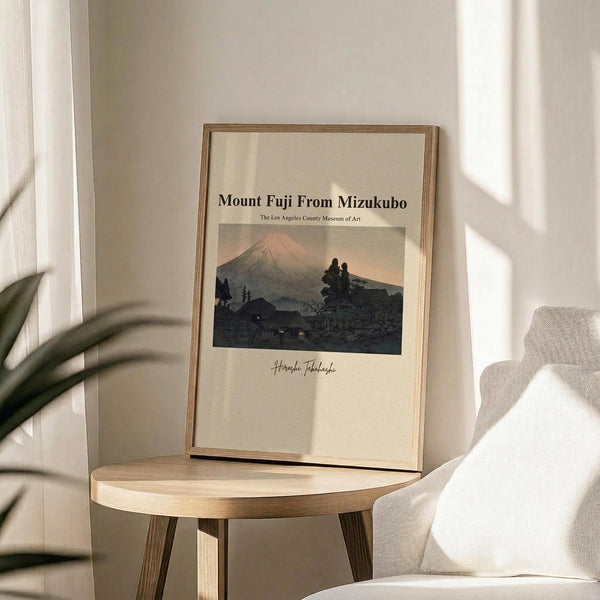 May include: Framed art print featuring Mount Fuji at sunset with traditional Japanese landscape in warm earth tones, displayed on light wood round side table next to beige upholstered chair. Natural sunlight creates soft shadows on neutral wall. Green plant leaves visible in corner. Thin natural wood frame surrounds cream-colored poster with mountain silhouette and small buildings in foreground.
