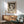 May include: Paul Klee exhibition poster featuring abstract figure with crescent moon in muted gray and cream tones, displayed in wooden frame above mid-century modern credenza. Scene shows sage green upholstered chair with wooden arms, white ceramic table lamp, and warm natural lighting casting shadows across white wall.