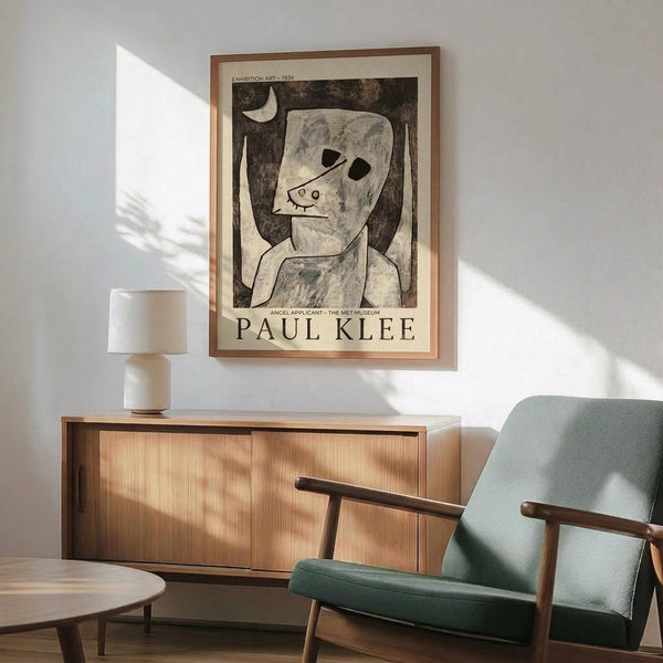 May include: Paul Klee exhibition poster featuring abstract figure with crescent moon in muted gray and cream tones, displayed in wooden frame above mid-century modern credenza. Scene shows sage green upholstered chair with wooden arms, white ceramic table lamp, and warm natural lighting casting shadows across white wall.