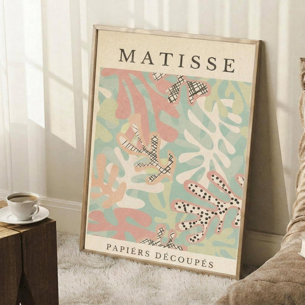May include: Framed Matisse-style art print with pastel pink, mint green, and cream abstract cut-out shapes and cross-hatched patterns, leaning against white wall corner. Wooden frame, positioned next to small wooden side table with white coffee cup and saucer, on textured cream rug with beige throw blanket visible.