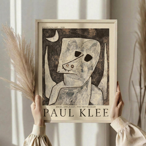 May include: Hands holding a framed Paul Klee exhibition poster featuring "Angel Applicant" artwork from 1939. The print shows an abstract figure with angular white form, dark geometric eyes, and simple line features against a textured brown background with crescent moon. Light wood frame, neutral beige clothing, dried pampas grass visible, soft natural lighting against white curtained window.