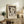 May include: Framed Paul Klee exhibition poster featuring abstract figure artwork in cream and brown tones, displayed on rustic wooden sideboard alongside potted fern, ceramic vase, and books in neutral-toned living room setting with beige upholstered furniture