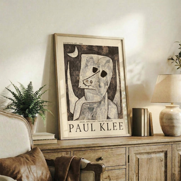 May include: Framed Paul Klee exhibition poster featuring abstract figure artwork in cream and brown tones, displayed on rustic wooden sideboard alongside potted fern, ceramic vase, and books in neutral-toned living room setting with beige upholstered furniture