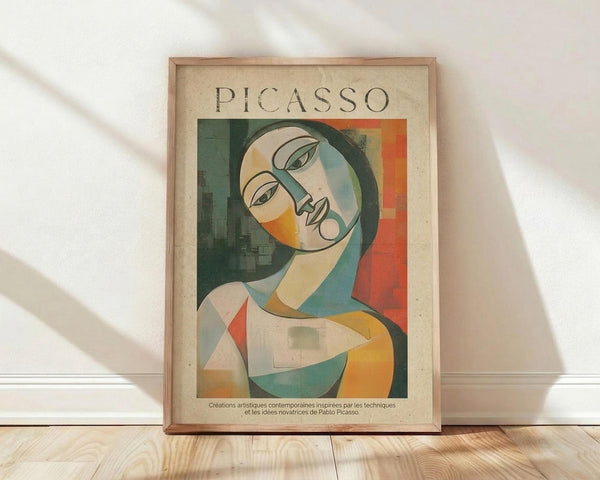 May include: Framed poster print featuring a cubist-style portrait of a woman with geometric facial features in green, orange, yellow and cream tones. The artwork shows "PICASSO" text at top with French description below. Natural wood frame sits against white wall on light wooden floor with soft window light casting shadows.