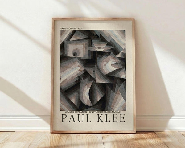 May include: Framed poster featuring Paul Klee's abstract geometric artwork from 1921 in muted grays, browns and soft tones with crystalline angular forms and gradated shading, displayed in light wood frame against white wall on natural wood flooring with soft natural lighting casting diagonal shadows across the scene.