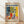 May include: Framed art print in natural wood frame leaning against white wall on light wooden floor. Poster features colorful still life with leafy plant branches, red ceramic vase, blue and white patterned pottery, bowl of yellow fruit on checkered tablecloth. Vibrant yellows, blues, reds in Matisse's distinctive cut-out style. Text reads "MATISSE" at top and "06200 NICE, FRANCE" at bottom.