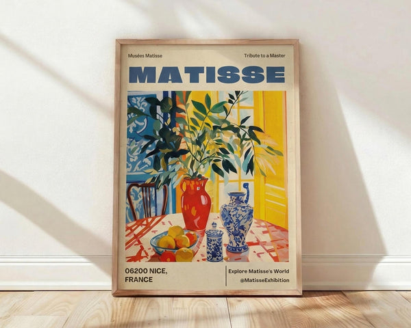 May include: Framed art print in natural wood frame leaning against white wall on light wooden floor. Poster features colorful still life with leafy plant branches, red ceramic vase, blue and white patterned pottery, bowl of yellow fruit on checkered tablecloth. Vibrant yellows, blues, reds in Matisse's distinctive cut-out style. Text reads "MATISSE" at top and "06200 NICE, FRANCE" at bottom.