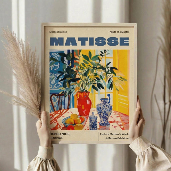 May include: Person in cream sweater holding framed museum poster featuring colorful still life with plants in red vase, blue and white ceramics, fruit bowl on patterned tablecloth. Artwork shows Matisse-inspired style with bold colors - yellows, blues, reds, greens. Light wood frame, dried pampas grass visible left side, soft natural lighting against cream curtained background.