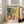 May include: Framed Matisse exhibition poster leaning against white wall next to beige sofa in bright living room. Colorful still life artwork shows red vase with green foliage, blue and white ceramic vessels, yellow fruit bowl on checkered tablecloth. Vibrant yellows, blues, reds in characteristic cut-out style. Natural light streams through windows onto hardwood floors.