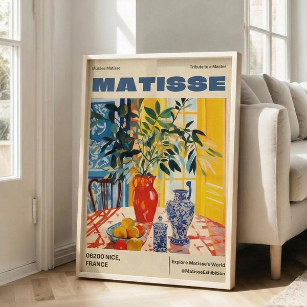 May include: Framed Matisse exhibition poster leaning against white wall next to beige sofa in bright living room. Colorful still life artwork shows red vase with green foliage, blue and white ceramic vessels, yellow fruit bowl on checkered tablecloth. Vibrant yellows, blues, reds in characteristic cut-out style. Natural light streams through windows onto hardwood floors.