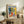 May include: Framed Matisse exhibition poster displayed on rustic wooden sideboard in neutral living room setting. Colorful artwork features still life with plants, vases, and fruit in blues, yellows, and reds. Scene includes fern plant, books, ceramic pottery, beige upholstered chair, and soft natural lighting creating warm shadows on cream walls.