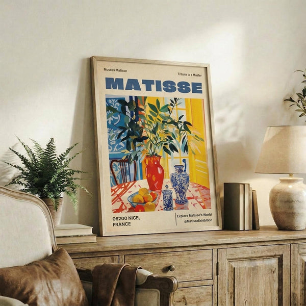 May include: Framed Matisse exhibition poster displayed on rustic wooden sideboard in neutral living room setting. Colorful artwork features still life with plants, vases, and fruit in blues, yellows, and reds. Scene includes fern plant, books, ceramic pottery, beige upholstered chair, and soft natural lighting creating warm shadows on cream walls.
