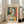 May include: Framed poster with abstract cubist-style woman's portrait in geometric shapes using green, orange, yellow and blue tones. Features "PICASSO" text at top and French text at bottom. White wooden frame leaning against white wall next to beige sectional sofa in bright living room with large windows and natural light casting shadows on hardwood floors.