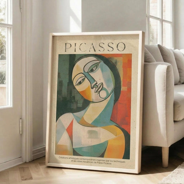 May include: Framed poster with abstract cubist-style woman's portrait in geometric shapes using green, orange, yellow and blue tones. Features "PICASSO" text at top and French text at bottom. White wooden frame leaning against white wall next to beige sectional sofa in bright living room with large windows and natural light casting shadows on hardwood floors.