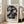 May include: Framed art print leaning against white wall next to beige upholstered chair in bright room with tall windows. Abstract geometric artwork features overlapping angular shapes in muted tones of gray, black, beige and sage green with crystalline fragmented pattern. Exhibition poster format with artist name at bottom in serif typography. Light wood frame, approximately 2-3 feet tall, positioned on hardwood flooring.
