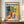 May include: Framed Matisse exhibition poster featuring vibrant still life with red vase, leafy branches, blue and white ceramics, and yellow fruit on checkered tablecloth, displayed on wooden console next to white ceramic vase and stacked books in elegant room with white paneled walls