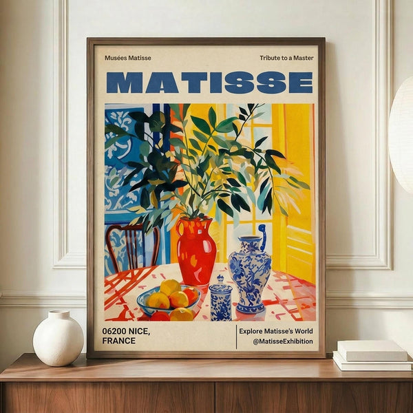 May include: Framed Matisse exhibition poster featuring vibrant still life with red vase, leafy branches, blue and white ceramics, and yellow fruit on checkered tablecloth, displayed on wooden console next to white ceramic vase and stacked books in elegant room with white paneled walls
