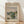 May include: Framed vintage-style exhibition poster featuring Claude Monet's impressionist landscape painting of a river scene with green foliage and soft cloudy sky, displayed against white wall on light wooden floor with natural window shadows