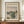May include: Framed exhibition poster featuring Claude Monet's impressionist river landscape with soft blues, greens, and peachy tones, displayed on a wooden sideboard beside a cream ceramic vase and white books against beige wainscoted walls.